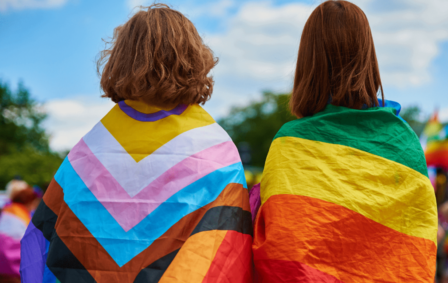 Two people stood in the sunshine with their backs turned carrying inclusive flags over their backs